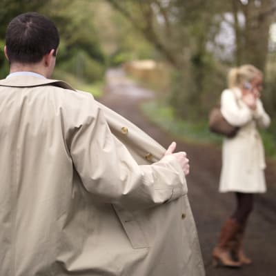 En man öppnar sin jacka framför en kvinna på en skogsväg.