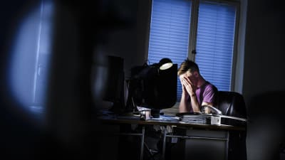 En person sitter i ett mörkt rum framför en dator. Han har huvudet i händerna och ser stressad ut.