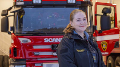 En kvinna står framför en brandbil och tittar in i kameran.