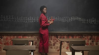 En rödklädd kvinna ritar vita streckgubbar på svarta tavlan i ett klassrum.