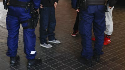 På bilden syns flera personers ben. På basis av kläderna kan man avgöra att det är två poliser som pratar med tre ungdomar.