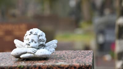 En vit kerubaktig ängel sitter med armarna i kors på en gravsten av marmor. 