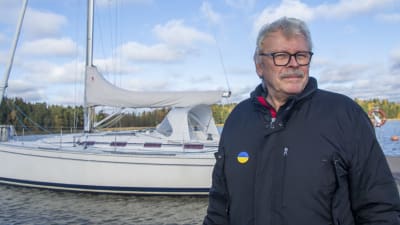 En man står på en brygga och tittar in i kameran. I bakgrunden skymtar en stor segelbåt.