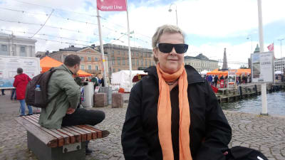 En kvinna i mörka solglasögon sitter vid kajen på Salutorget i Helsingfors. 