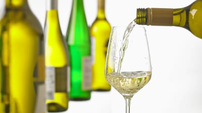 Vitvin hälls upp i ett glas, en rad vitvinsflaskor i bakgrunden.