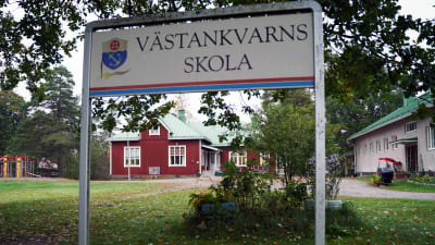 En röd, äldre byskola skymtar bakom en skylt där det står Västankvarns skola.