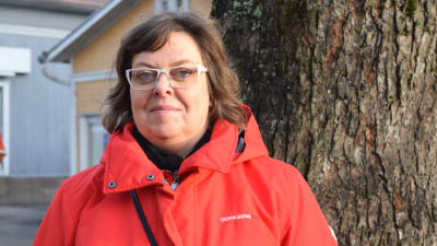 En kvinna med röd jacka och glasögon poserar ute vid ett större träd inne i en stad.