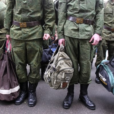 Joukko uusia varusmiehiä seisoo rivissä armeijan univormuissa, käsissään siviilireppuja ja laukkuja. Kuva on rajattu niin, että varusmiesten kasvoja ei näy.