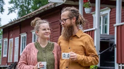 Ruut Luukkonen och Jakob Tjäder står vid ett gammalt rött trähus och dricker kaffe. 