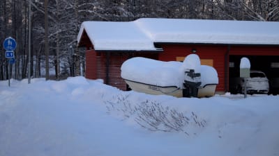 En fritidsbåt med utombordsmotor står ute på egnahemsgård, allt helt täckt av snö.