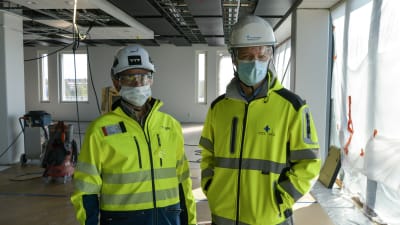 Två män i arbetskläder, hjälm, skyddsglasögon och munskydd står i ett stort rum som är under konstruktion.