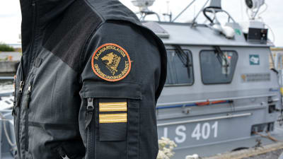 Anonym person i svart jacka. Gränsbevakningens emblem syns på axeln. En patrullbåt finns i bakgrunden.