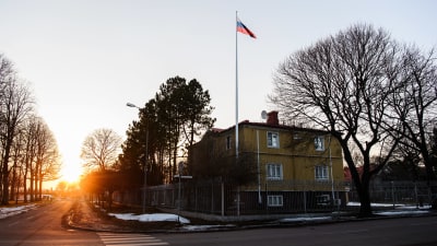 Rysslands konsulat i Mariehamn en vårdag. Det finns lite snö på marken utanför trevåningshuset vars översta två våningar är gula. En rysk flagga vajar i en flaggstång och på taket till byggnaden syns en satellitantenn.