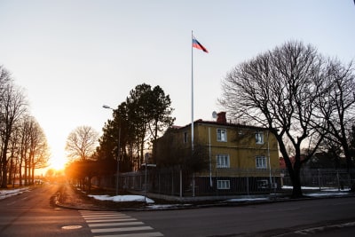 Rysslands konsulat i Mariehamn en vårdag. Det finns lite snö på marken utanför trevåningshuset vars översta två våningar är gula. En rysk flagga vajar i en flaggstång och på taket till byggnaden syns en satellitantenn.
