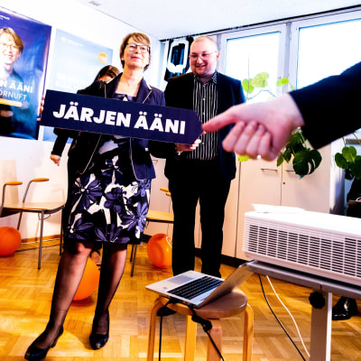 Sari Essayah järjen ääni vaalikyltti kädessään vaalivalvojaissa. Taustalla ehdokkaana ollut Kauko Pohjanen.