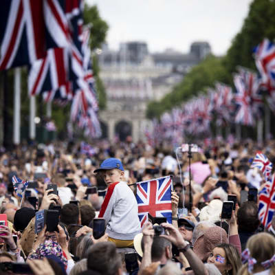 Suuri ihmisjoukko on kokoontunut Iso-Britannian lippujen reunustamalle tielle kuvaamaan ja katselemaan. Lapsi istuu aikuisen olkapäillä ja kääntyy katsomaan kameraan.