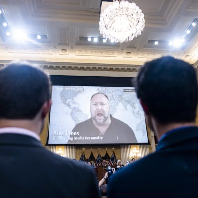 Alex Jones visas på en storskärm i en sal med människor. Under skärmen syns amerikanska flaggor.