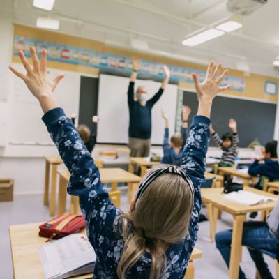 Barn i ett klassrum räcker upp sina armar i luften.