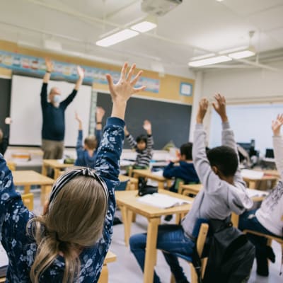 Barn i ett klassrum räcker upp sina armar i luften.