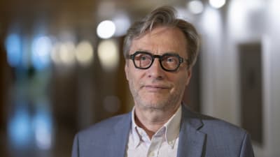 En närbild på ledande överlekare vid Helsingfors och Nylands sjukvårdsdistrikt, Markku Mäkijärvi. Mäkijärvi har glasögon med svarta bågar och en grå kostym på sig. 