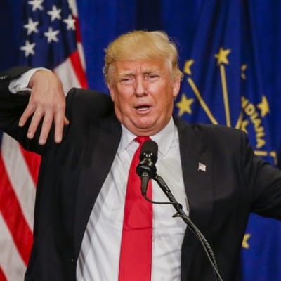 Donald Trump viittilöi puhuessaan mikrofoniin. Hänellä on tumma puku, valkoinen kauluspaita ja punainen kravatti. Takin rintapielessä on Yhdysvaltain lippua esittävä pinssi. Taustalla näkyy Yhdysvaltain lippuja ja Indianan osavaltion lippuja.