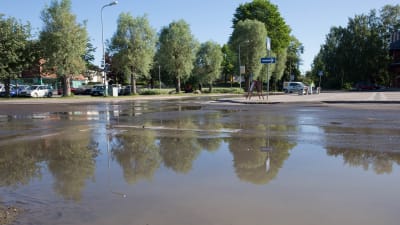 Ett vattenläckage har orsakat stora mängder vatten på en asfalterad väg. I bakgrunden skymtar en byggnad och några träd.