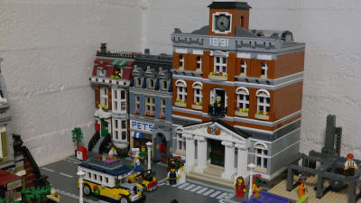 En bit av en stad byggd av legobitar. En byggnad ser ut som ett rådhus. På gatan finns en gammaldags buss av lego.
