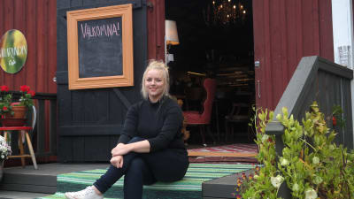 Ellen Järvinen sitter på trappan framför en öppen dörr till till sitt café Källarvinden i Kasnäs
