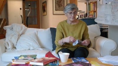 Margita Vainio sitter i en soffa med en hel hop med böcker framför sig