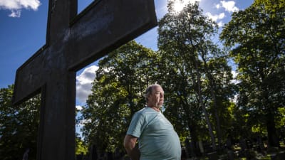 En man står med händerna bakom ryggen framför ett stort kors på en kyrkogård.