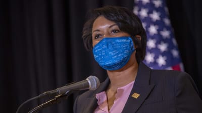 En kvinna i kritstrecksrutig kavaj och ljusrosa skjorta talar i en mikrofon. Hon har på sig ett blått munskydd. Bakom henne syns en amerikansk flagga.