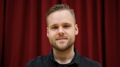 Mansperson står på teaterscen, i bakgrunden rött sammetsdraperi.