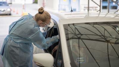 Närvårdare i skyddsdräkt tar coronaprov av klient sittande i bil. 