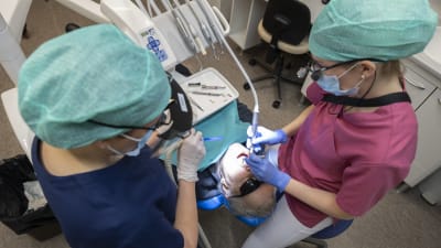 Två personer sköter om tänderna på en äldre man i tandläkarstol.