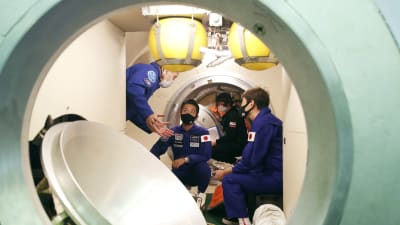 Tre personer i blå overaller och munskydd inne i ett rymdskepp.