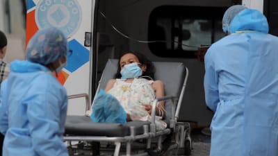 En kvinna i munskydd ligger på en bår medan hon lassas in i en ambulans av två sjukskötare i skyddsutrustning.