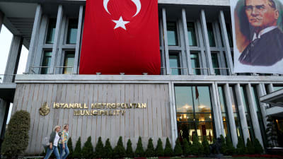 En bild på människor som går förbi ett hus. Turkiet flagga hänger på huset. 