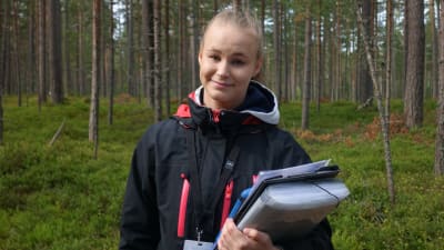 Janni Heikkinen med svart jacka, namnskylt och mappar i handen. En tallskog i bakgrunden.