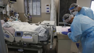 Coronapatient som ligger i en sjukhussäng och två läkare eller sjukvårdare som står framför sängen och antecknar något på ett papper.