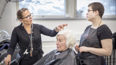 En frisörslärare och en studerande står på var sin sida av en kund och funderar på hennes frisyr. 