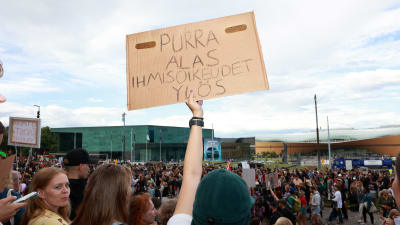 Demonstranter utanför riksdagshuset med skylt som kritiserar Riikka Purra.