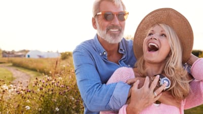 Äldre man i solglasögon omfamnar leende yngre kvinna i solhatt