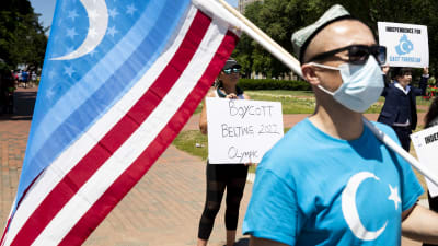En man i munskydd och solglasögon bär både den amerikanska och uiguriska flaggan över axeln. Bakom honom syns en kvinna med ett plakat med texten "Boycott Beijing 2022 Olympic".