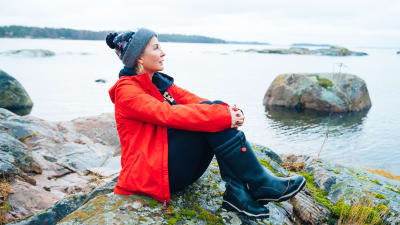 Kvinna i röd jacka sitter på en klippa och ser ut över havet.