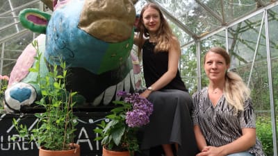 Två kvinnor sitter intill en konstnärlig och färgrann koskulptur inne i ett växthus.