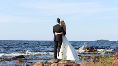 Bröllopsklätt par står och ser ut över havet med ryggen mot kameran