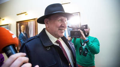 En äldre man i hatt blir fotograferad och har mikrofoner framför sig.