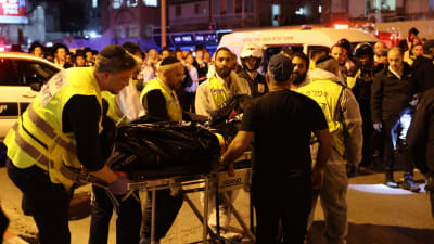 Polis rullar på en bår med en svart liksäck på. I bakgrunden en ambulans.
