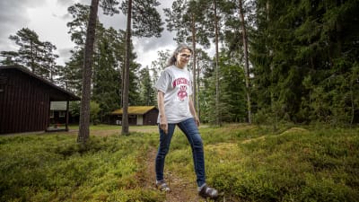 En kvinna går utomhus längs en stig. I bakgrunden syns två byggnader.