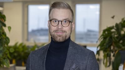 En man med glasögon, tröja och kavaj ser in i kameran.